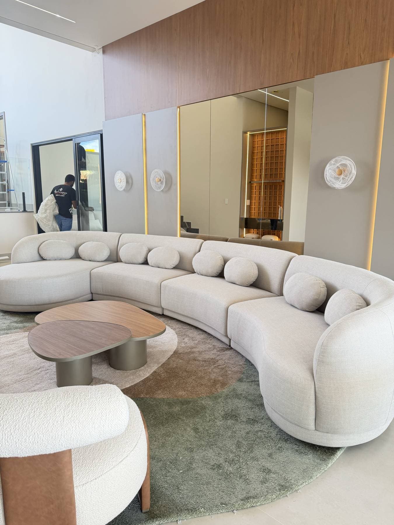 Sala de estar com sofá curvo claro, mesas de centro de freijó e painel de madeira ao fundo